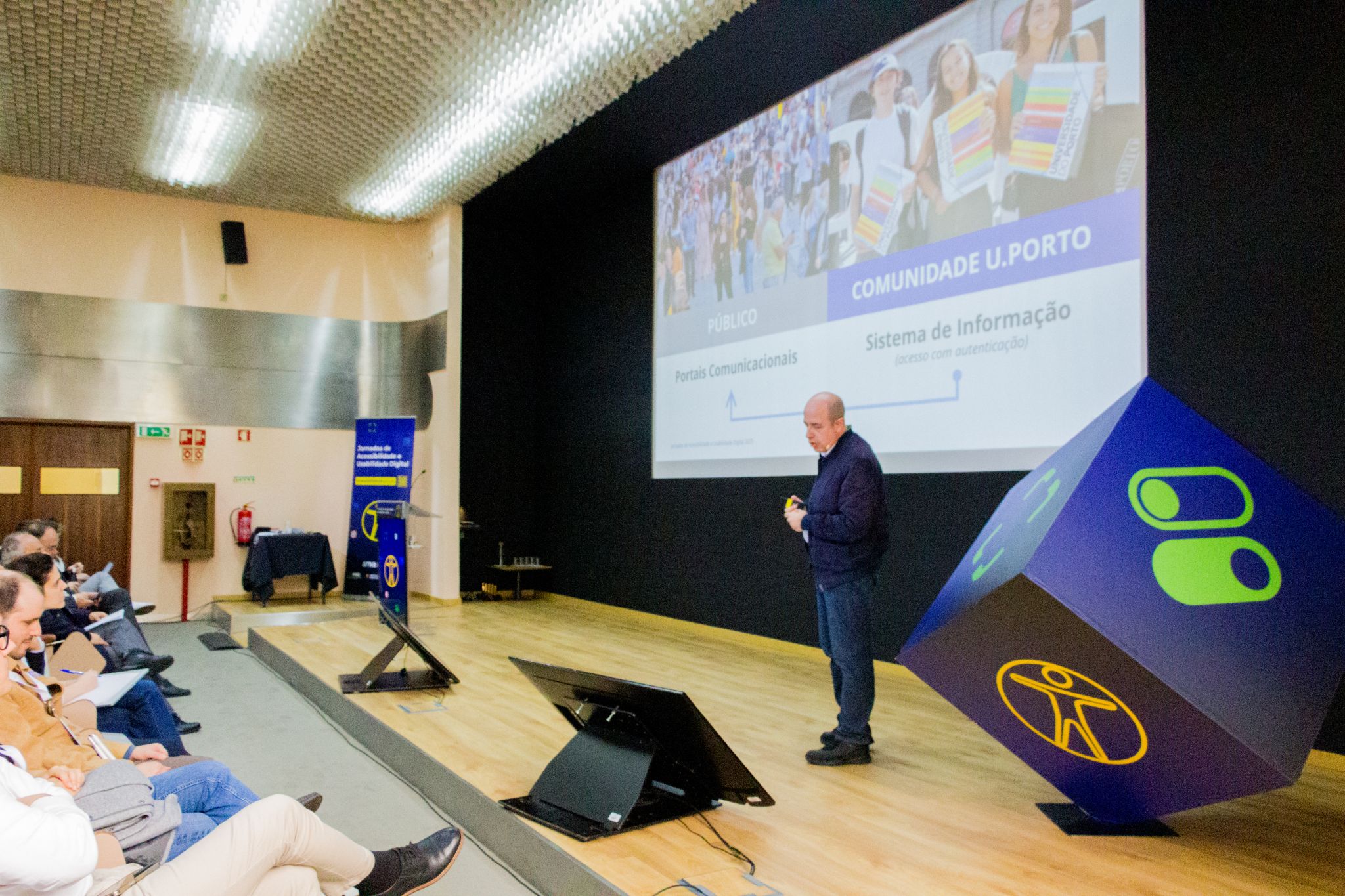 Sessão plenária no auditório com orador em palco a apresentar um caso prático, perante uma audiência atenta.